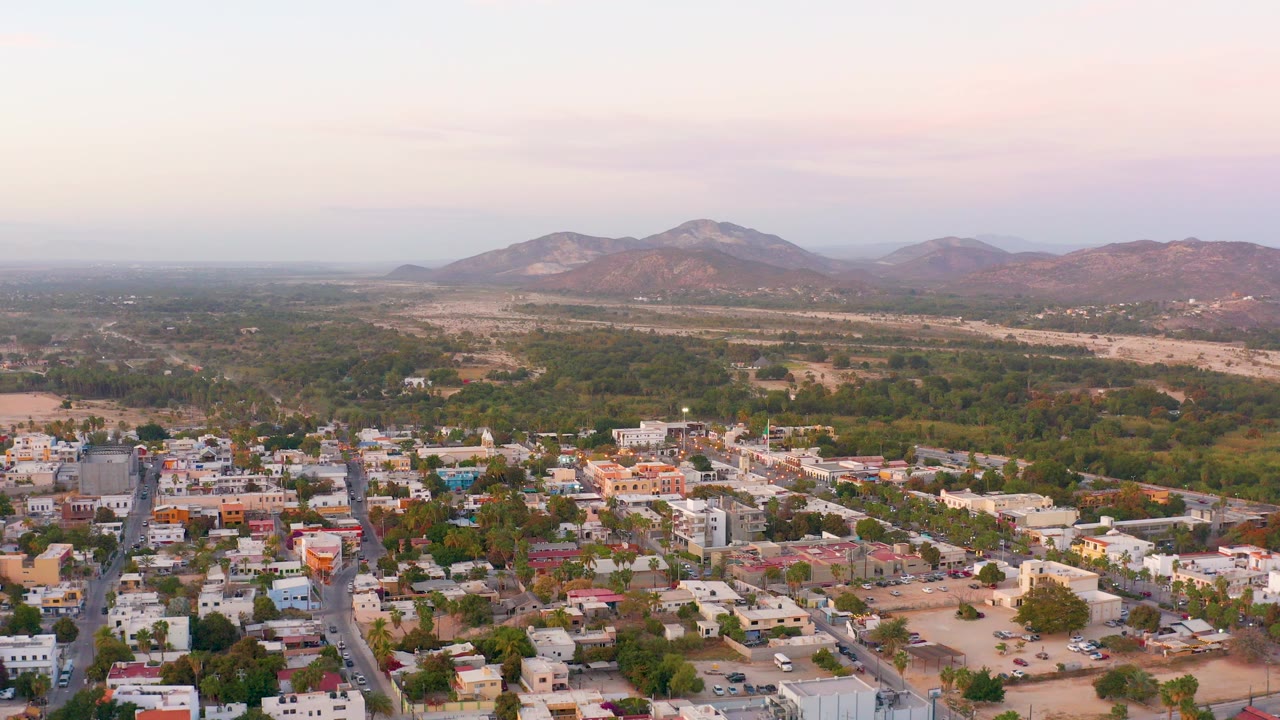 Opening aerial frame over San Jose del Cabo, with residential buildings and streets in the lower frame, green areas and dry ground beyond, mountains on the horizon, and no visible people.