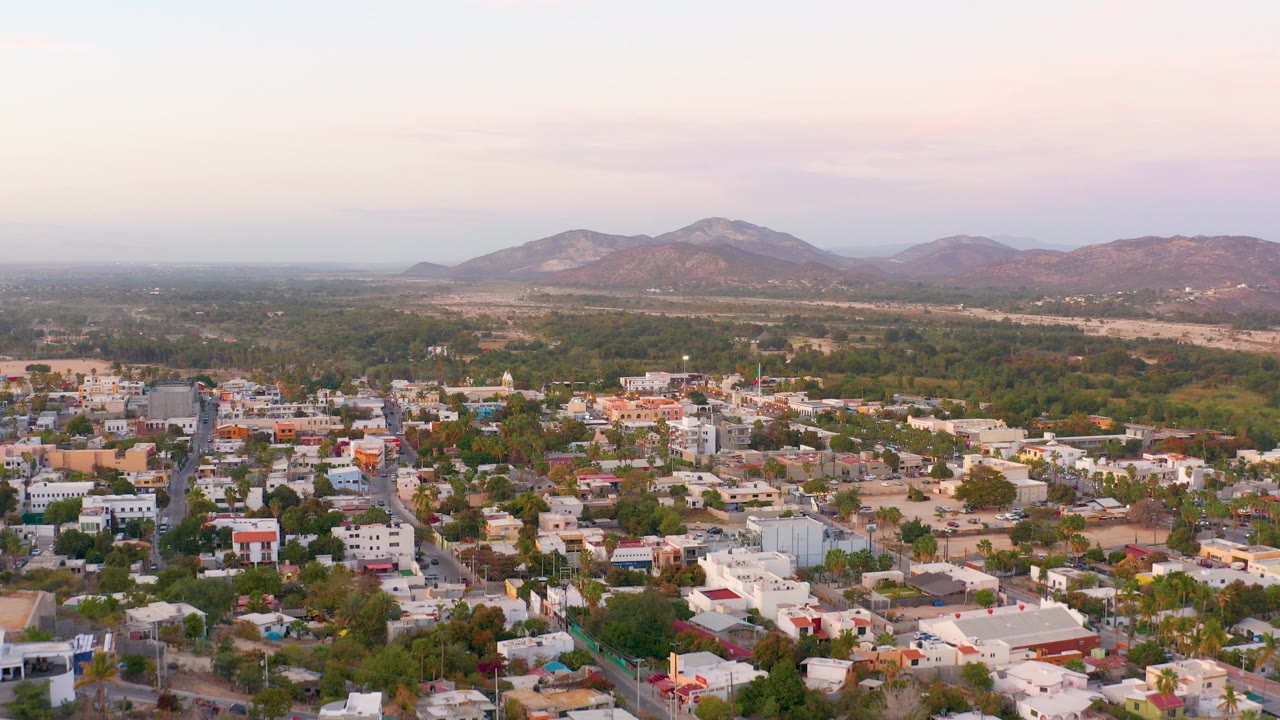 Aerial view over San Jose Del Cabo homes and gridded streets, with trees, open land, distant mountains, a pale pink sky, and no visible people.