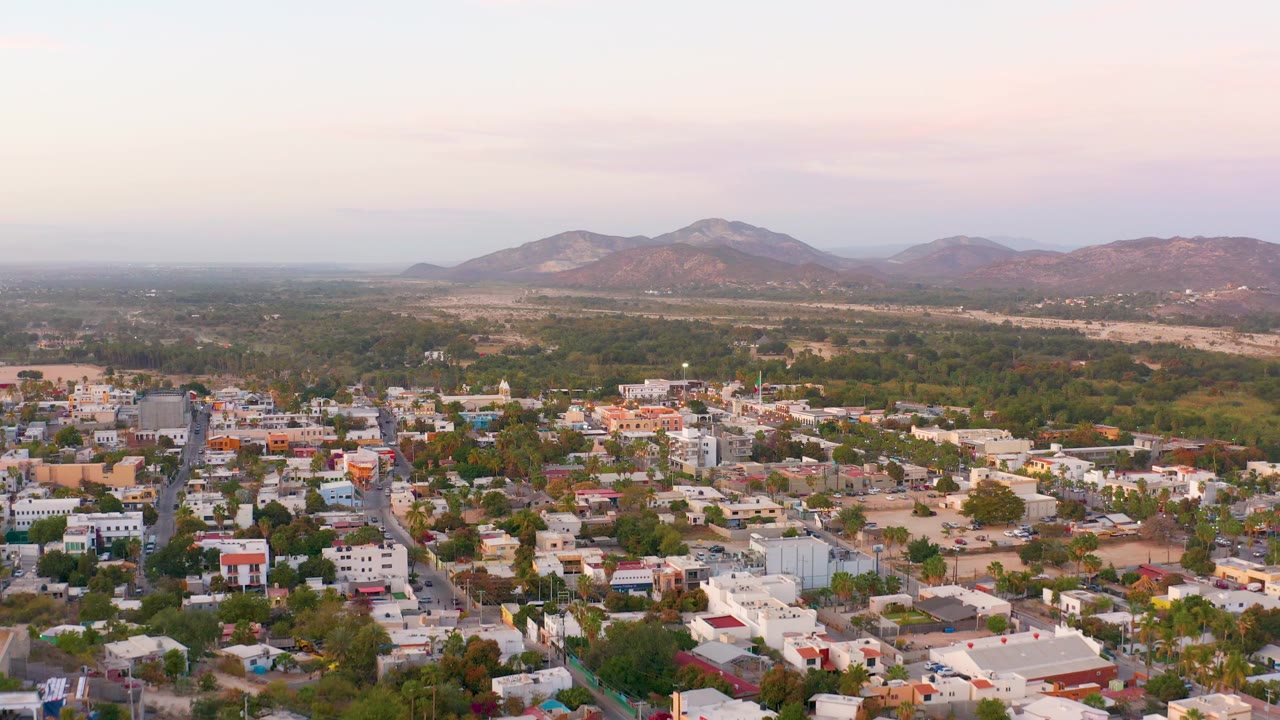 Wide aerial view of San Jose Del Cabo rooftops and streets in soft light, with green midground terrain, low hills, larger mountains, and no visible people.