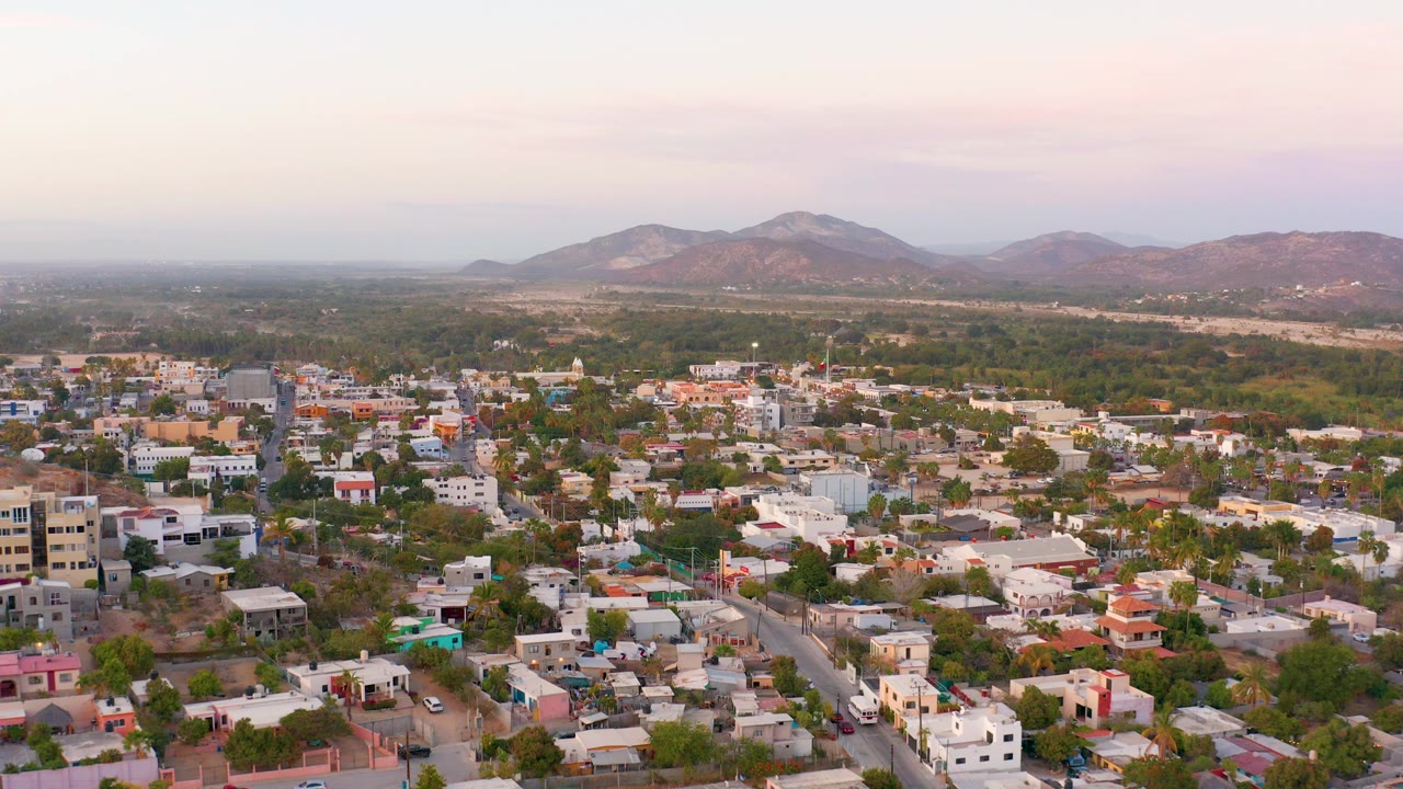 Aerial view over San Jose Del Cabo residential blocks, scattered trees, pale streets, and distant mountains under a soft pink sky, with no visible people.