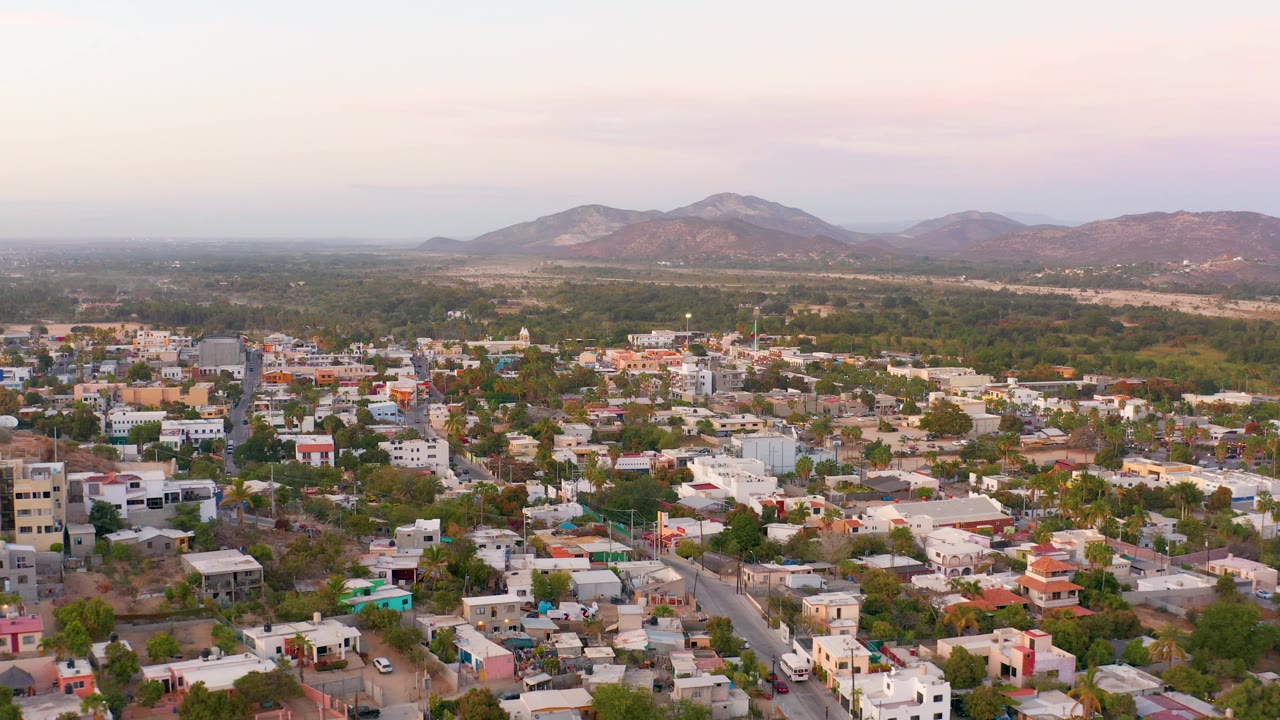 Wide aerial frame over San Jose Del Cabo rooftops and road corridors, with mixed trees, open land, and brown-green mountains under pastel cloud cover, with no visible people.