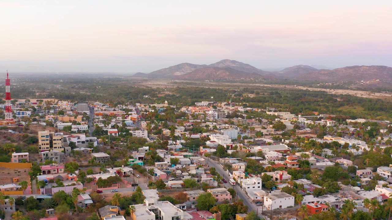 Aerial view of San Jose Del Cabo with the tower closer to the left edge, dense low-rise rooftops in the foreground, green midground, distant mountains, and no visible people.