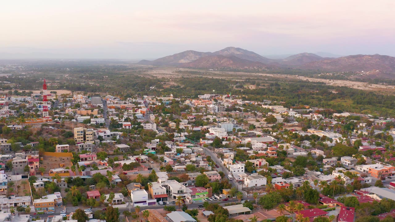 San Jose Del Cabo residential area seen from above, with the red and white tower nearer the center, rows of homes, green trees, mountains, and no visible people.