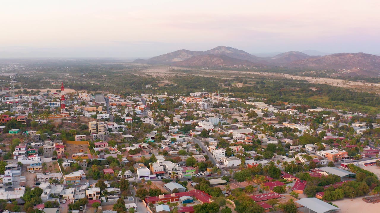 Opening frame over San Jose Del Cabo residential blocks, with pale rooftops, green trees, dry hills, distant mountains, soft pink sky, and no visible people.