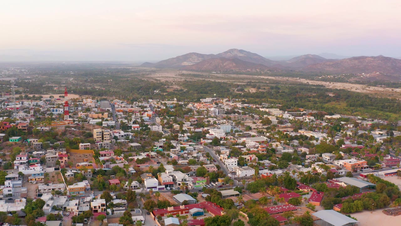Second frame shows San Jose Del Cabo rooftops and tree cover slightly farther across the neighborhood, with the mountain ridge clear in the background and no visible people.
