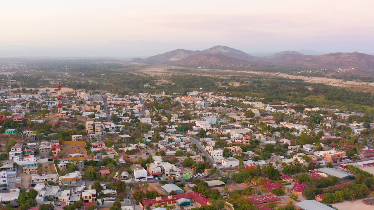 Third frame holds on San Jose Del Cabo houses, palms, and scrubland, with dry slopes and distant mountains across the upper half of the image and no visible people.