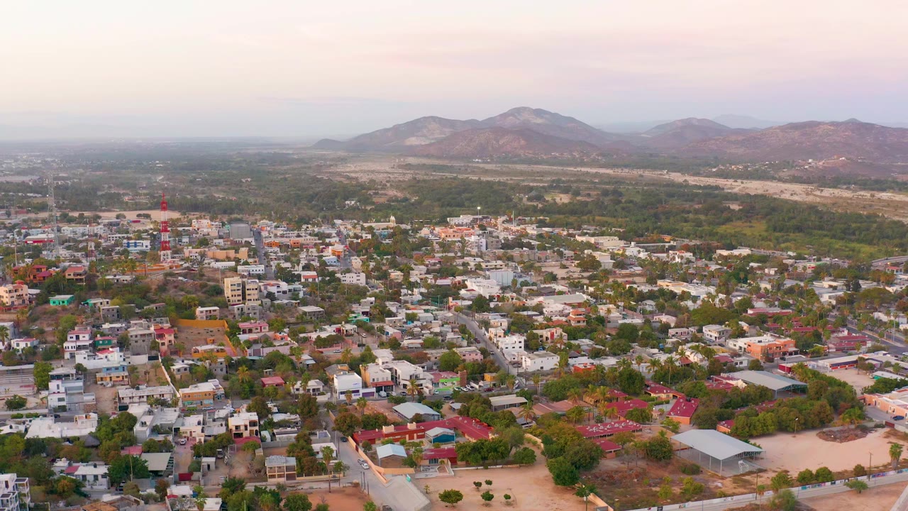 Early aerial frame of San Jose del Cabo with clustered houses, green tree cover, open tan ground, a red tower, mountain ridges, and no visible people.