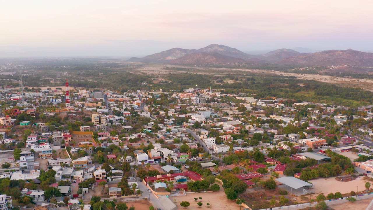 Aerial view over San Jose del Cabo residential blocks, with white and pastel buildings in the foreground, a green midground belt, hazy hills, and no visible people.