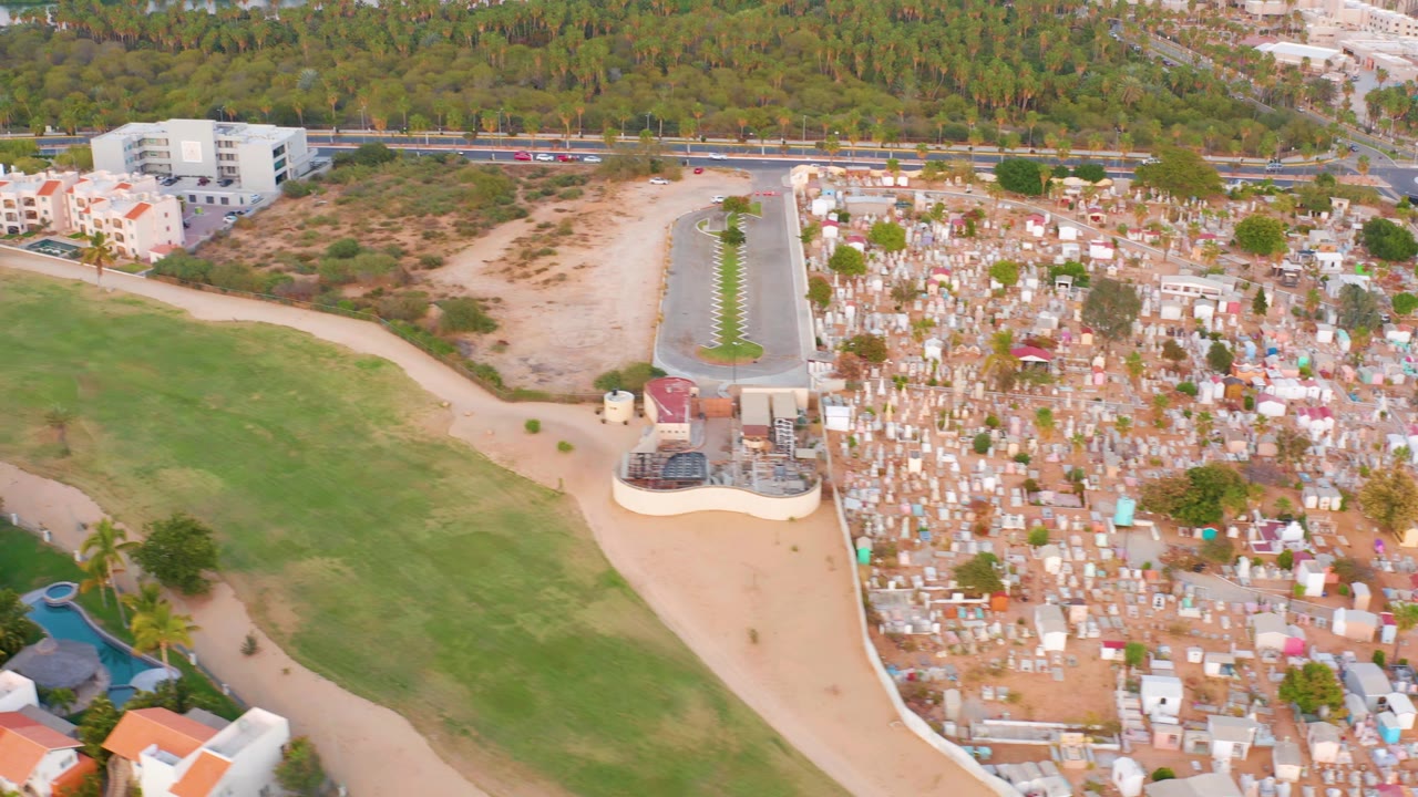 Drone view of cemetery edge, open grass, road, residences, trees, and water.