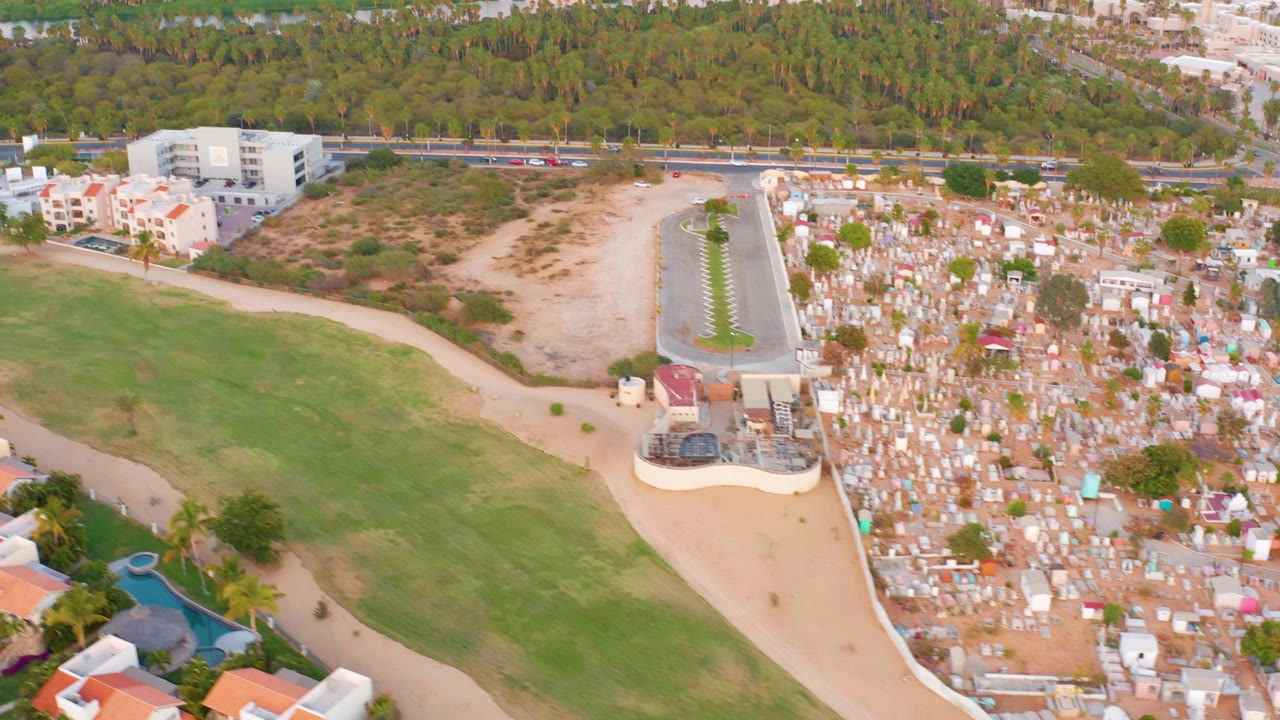 San Jose del Cabo aerial frame with cemetery markers at right, sandy open land and green field at center, a roadway with small vehicles, white buildings, dense trees, and a water channel behind them, no visible people.