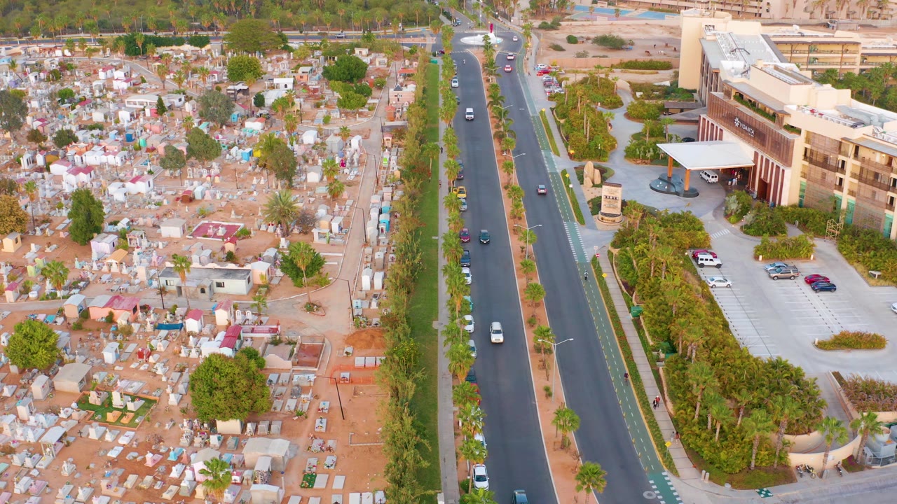 Aerial view in San Jose del Cabo, Mexico, showing a divided road between dense small structures on reddish ground and a larger building with a parking lot, with no visible people.