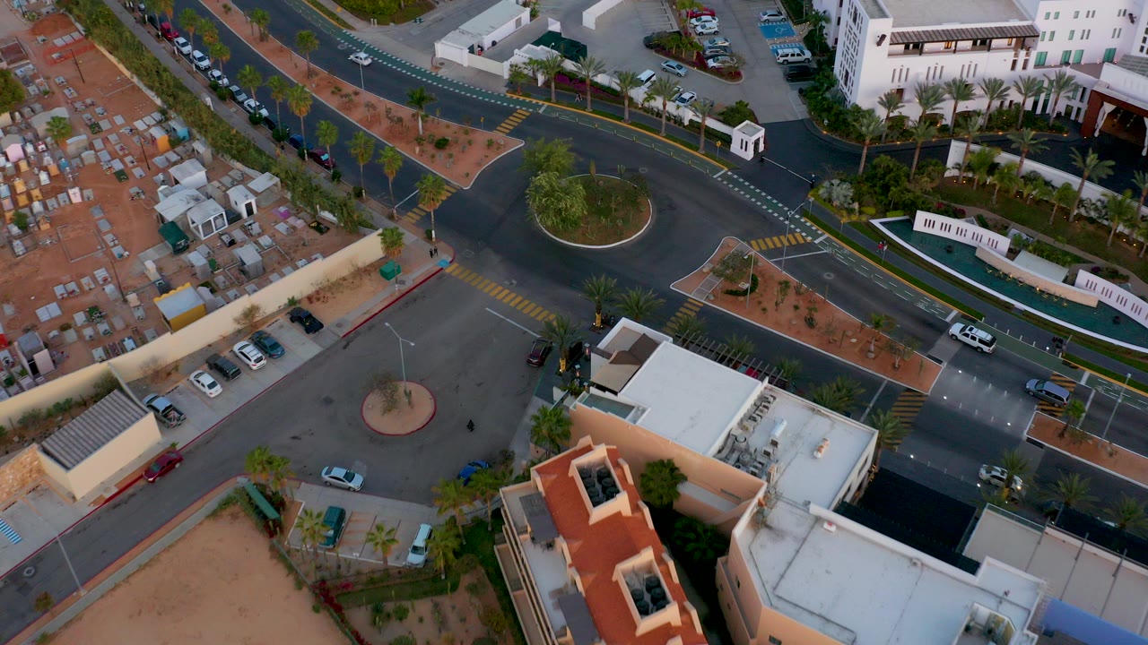 Overhead frame in San Jose Del Cabo showing a roundabout, dark roads, yellow crosswalks, parked cars, palms, and low buildings with no clearly visible people.