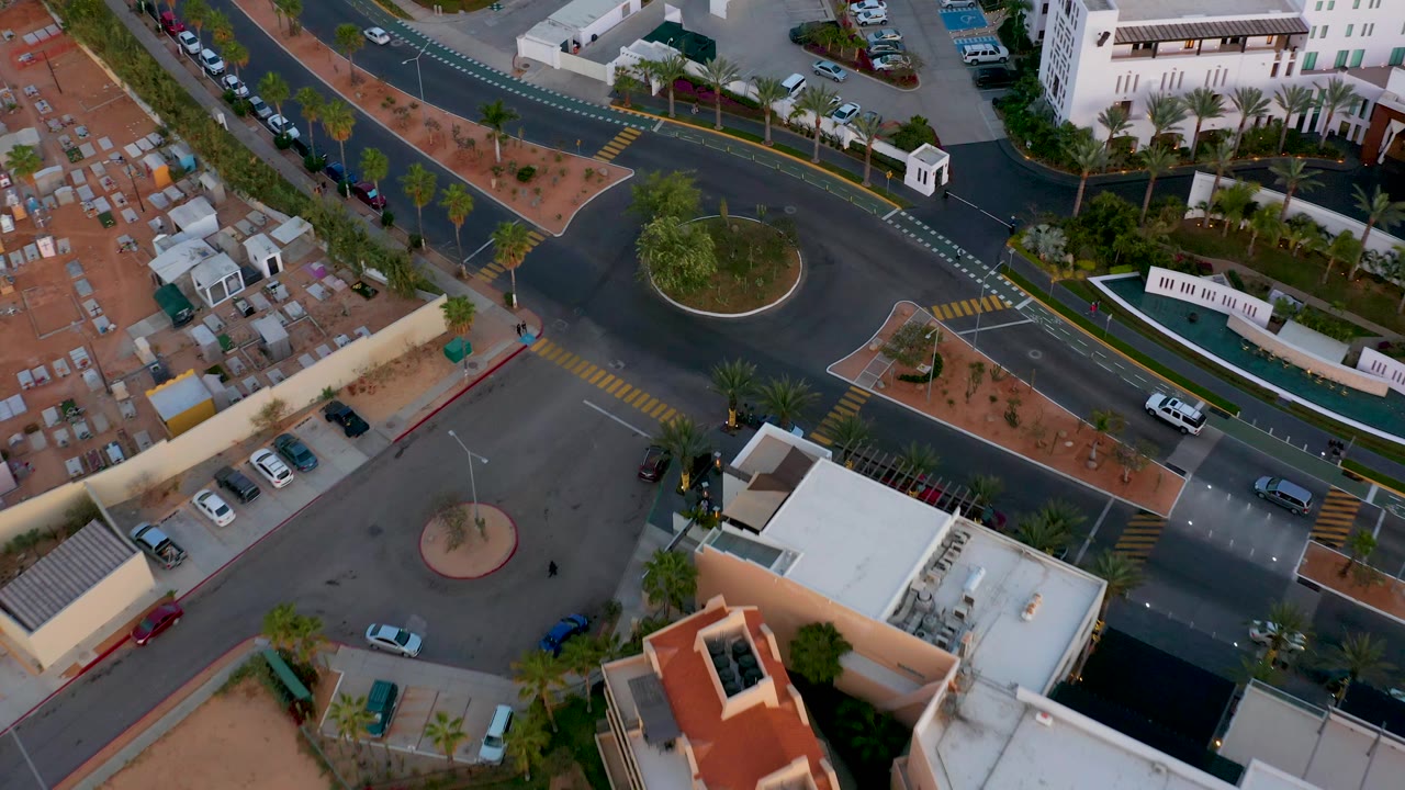 Top-down San Jose Del Cabo frame showing the roundabout, a broad paved parking area, surrounding roads, parked cars, a white building, and no clearly visible people.
