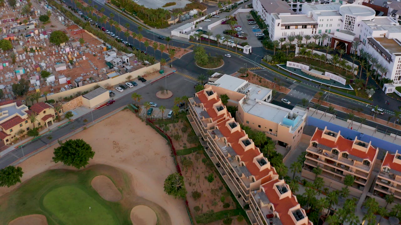 Opening aerial frame over San Jose del Cabo showing a golf green and sand traps below red-roof residential buildings, roads, parked cars, and cemetery plots, with no visible people.