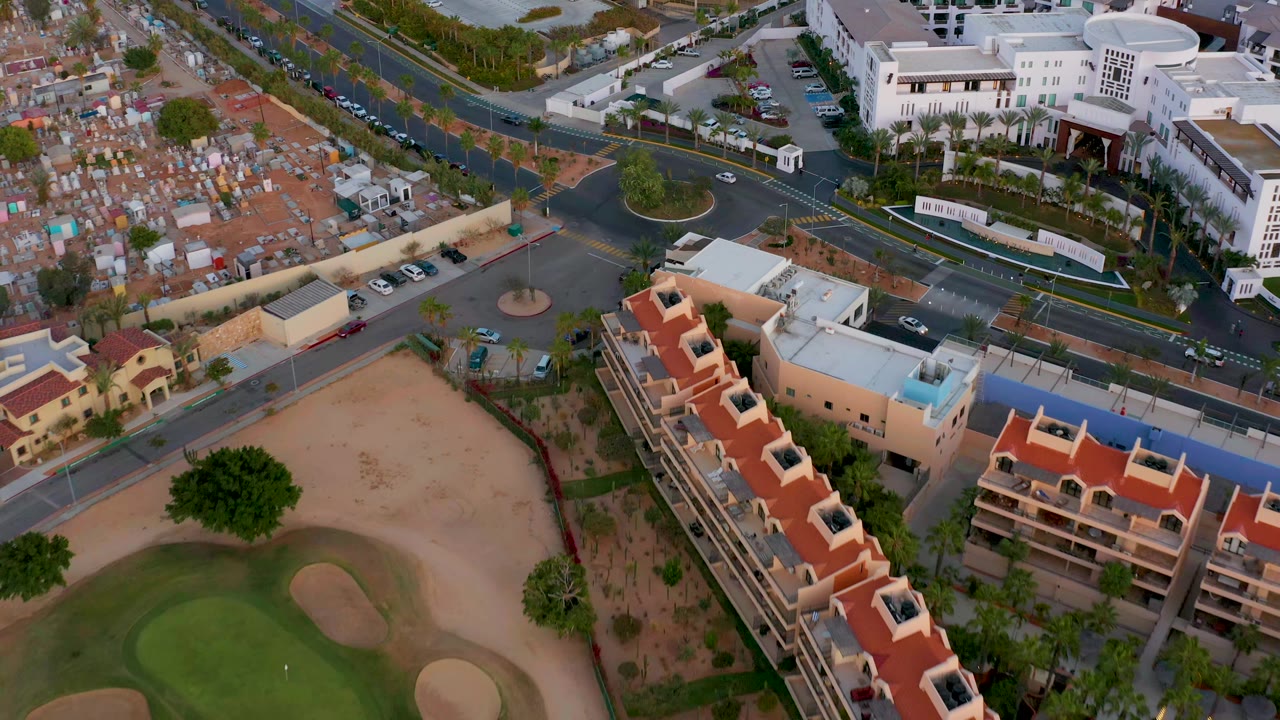 Drone view of residential buildings beside a golf course and roads near cemetery plots.