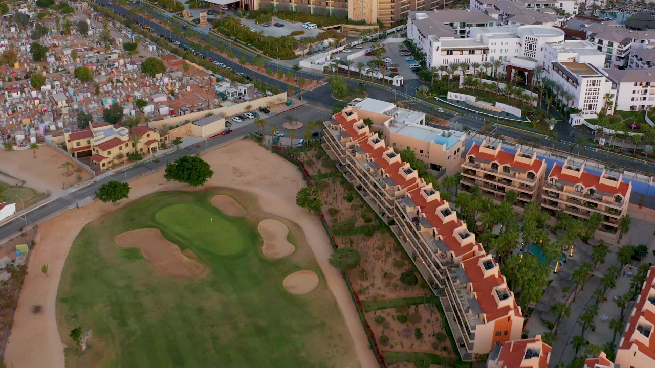 Aerial view in San Jose Del Cabo, Mexico with the golf course centered lower frame, apartment buildings to the right, a road intersection above, and no visible people.