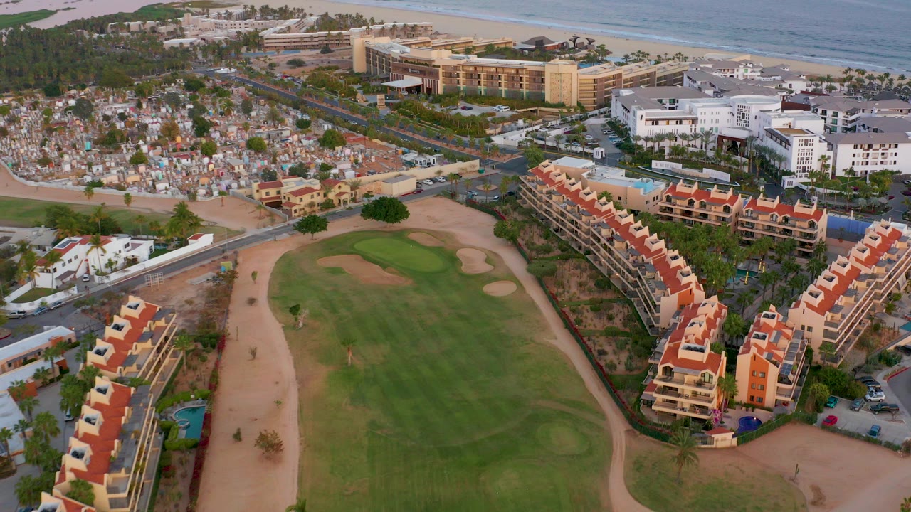 Aerial frame over San Jose del Cabo showing a green golf fairway, sand traps, red-roof residential buildings, white buildings, beach, ocean, and no visible people.
