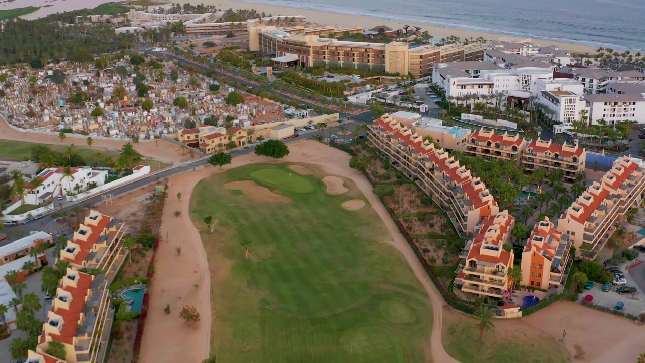 Aerial frame in San Jose del Cabo with the golf green centered lower in the image, red-roof residences to the right, town blocks to the left, ocean at the top, and no visible people.