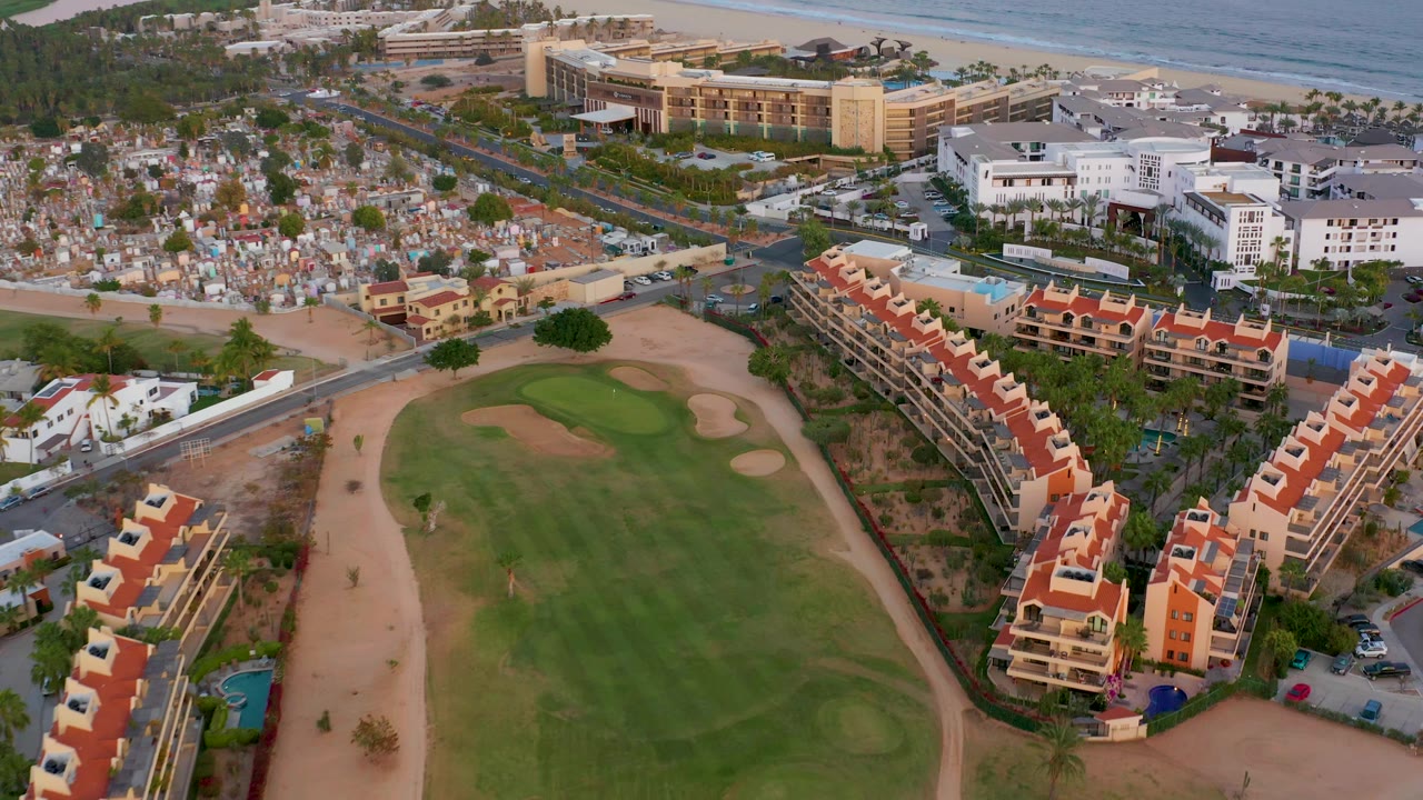 Aerial frame over San Jose del Cabo showing more of the right-side red-roof residential rows, the central golf fairway and bunkers, white buildings near the beach, ocean, and no visible people.