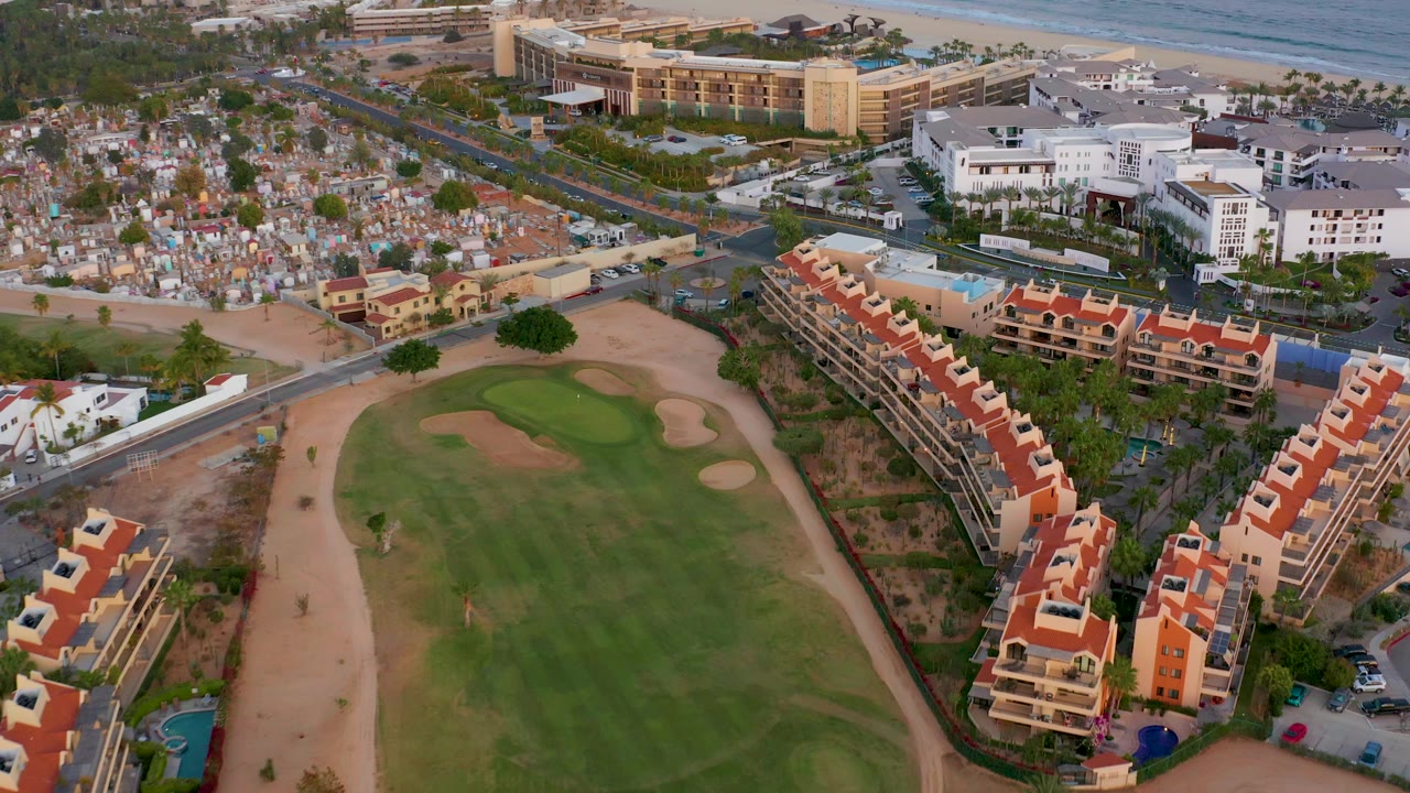 Aerial view across a golf course toward town blocks, white buildings, beach, and ocean.