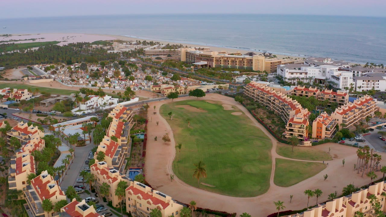 Opening aerial frame over San Jose Del Cabo showing a green golf course, red-roofed resort buildings, white hotel blocks, beach sand, and the ocean horizon with no visible people.