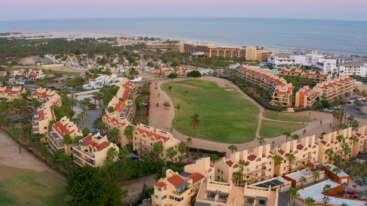 Early frame of San Jose Del Cabo showing red-roof residential buildings around a large green lawn, with beach and ocean in the background.