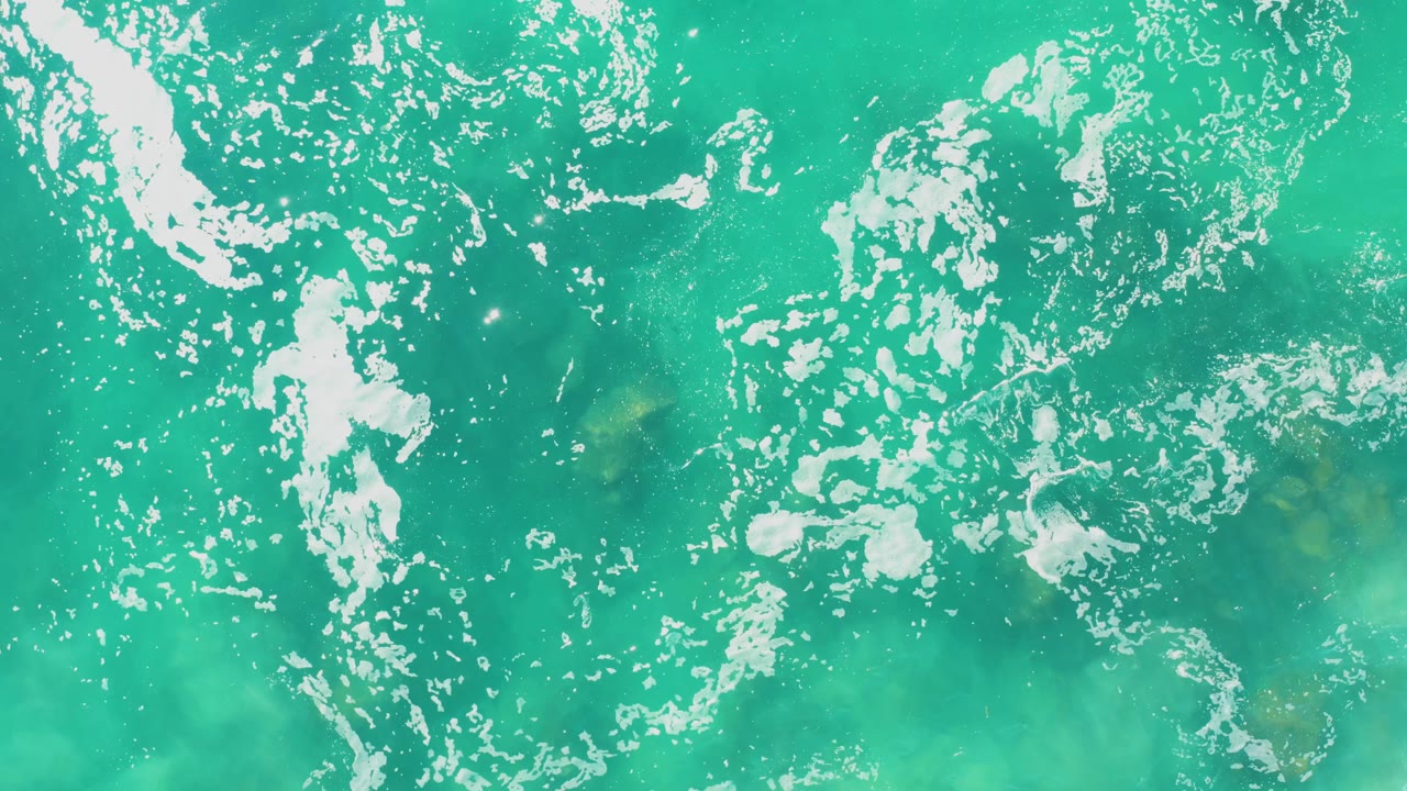 Turquoise water at Laguna Beach Los Cabos shows scattered white foam over pale submerged rocks, with no visible people.
