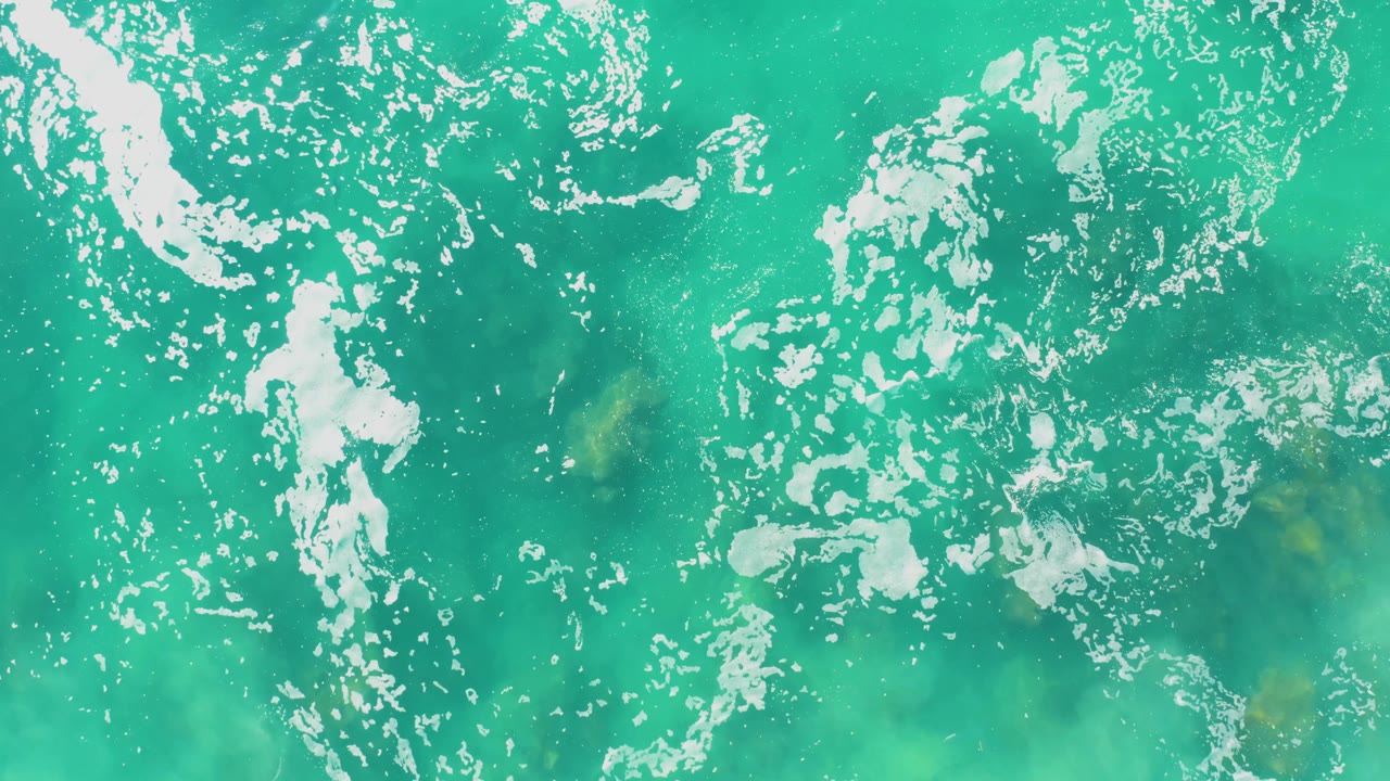 Clear green-blue water fills the frame at Laguna Beach Los Cabos, with broken foam streaks and submerged rock shapes visible below, and no visible people.