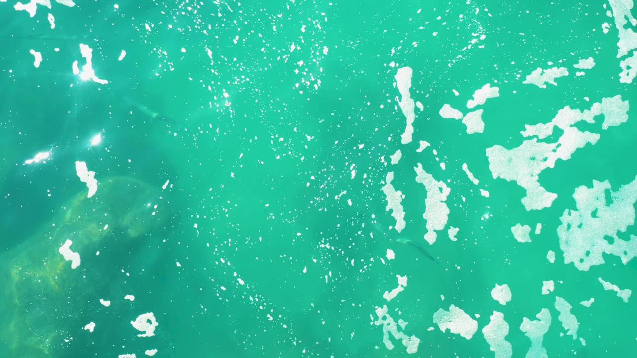 Top-down view of turquoise shallow water at Laguna Beach Los Cabos with scattered white foam, faint submerged rocks, and no visible people.