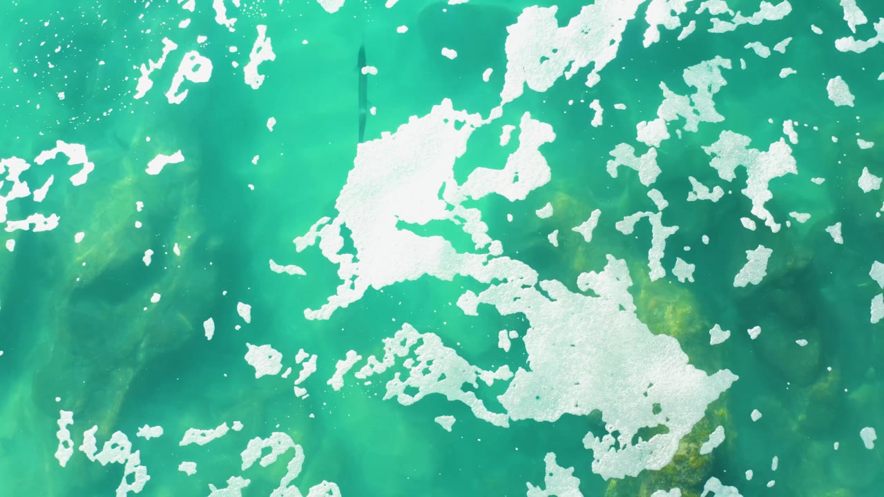 Top-down view of Laguna Beach Los Cabos water with broken white foam fields spread across bright turquoise and green surface tones, with no visible people.