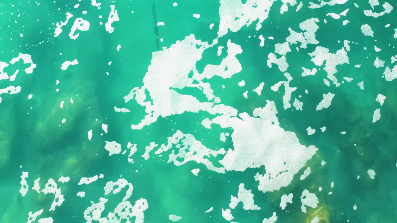 Aerial-style view of Laguna Beach Los Cabos ocean with a larger white foam mass left of center and darker green water visible beneath, with no visible people.