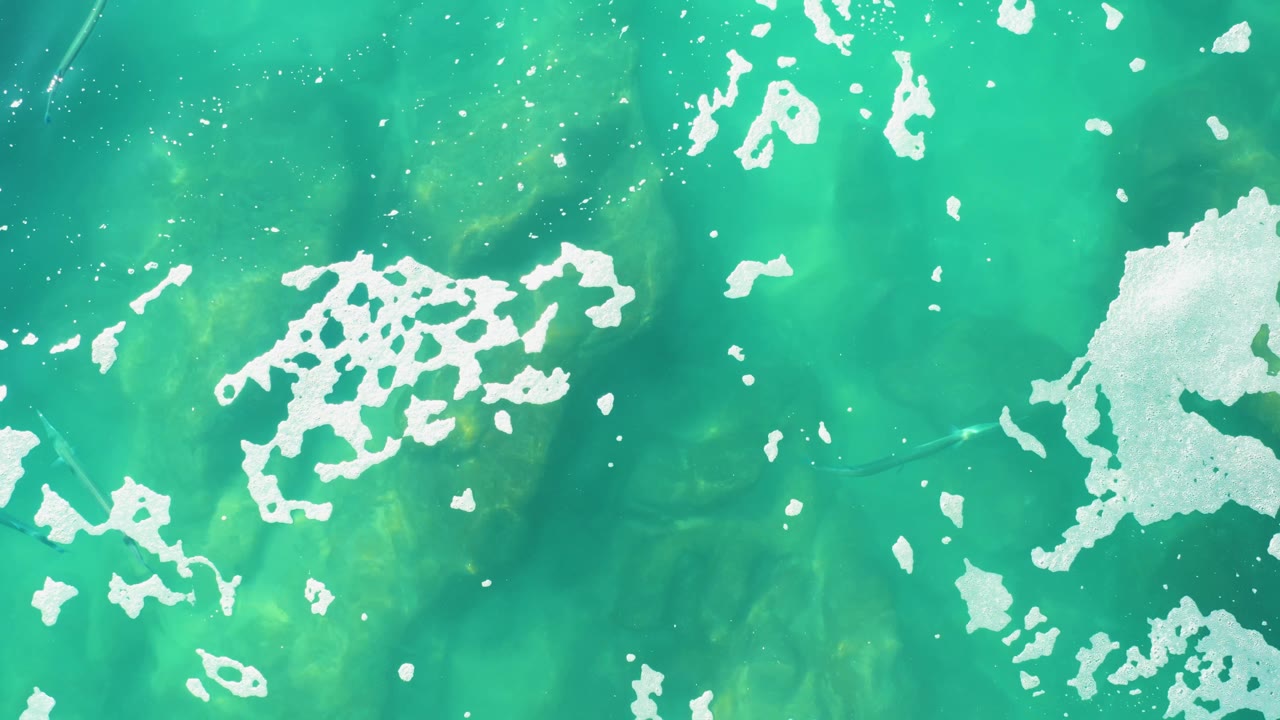 Clear turquoise ocean at Laguna Beach Los Cabos seen from above, with bright foam patches spread across the upper and side areas and no visible people.
