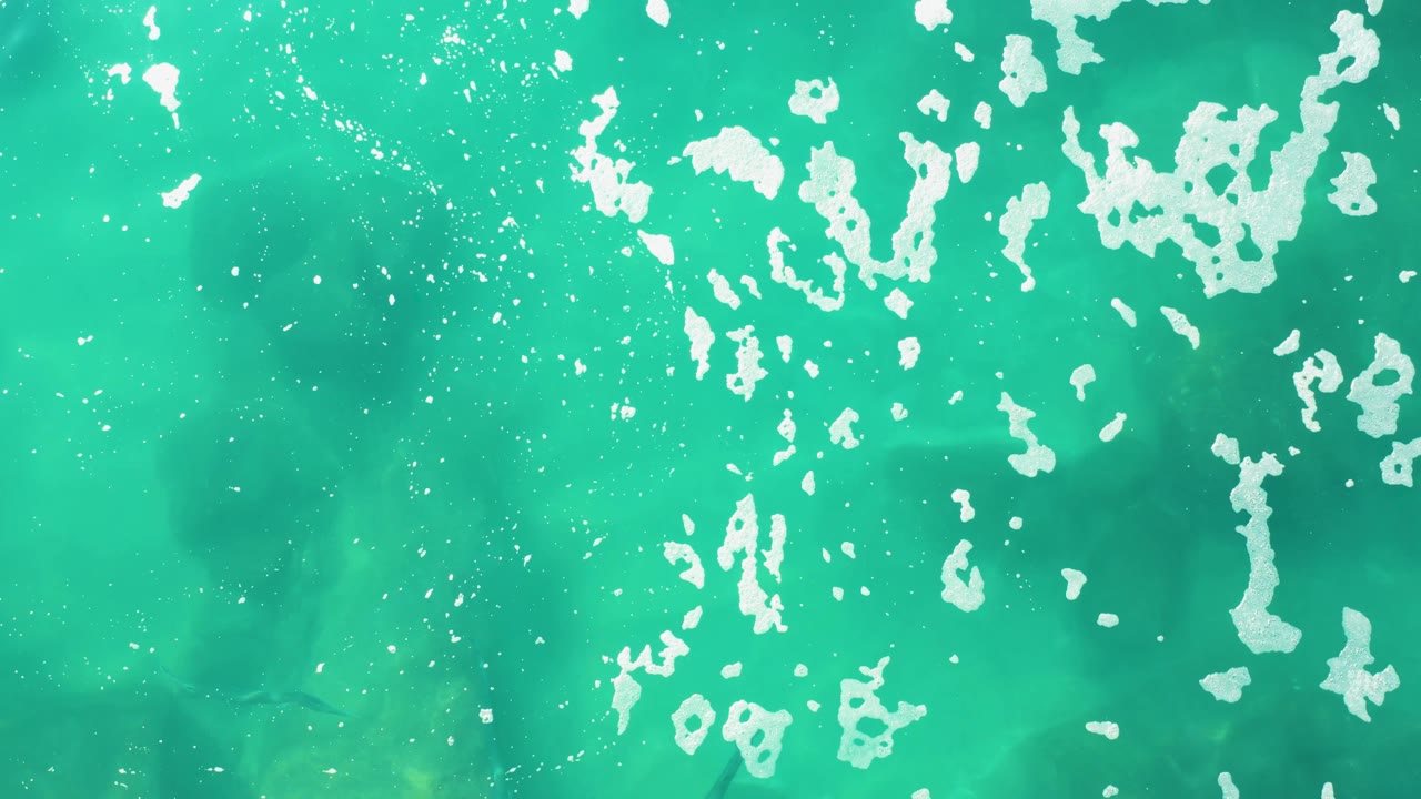 An overhead frame from Laguna Beach Los Cabos shows bright turquoise water, broken white foam flecks, faint underwater bottom texture, and no visible people.