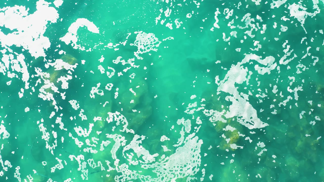 Overhead frame of Laguna Beach Los Cabos water where bright white foam clusters sit over green turquoise ocean, no visible people.
