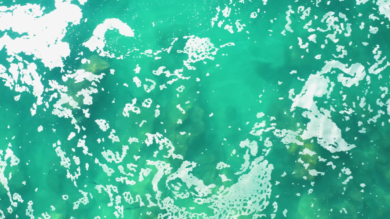 Overhead turquoise sea surface at Laguna Beach Los Cabos with scattered bubbles, irregular white foam, and faint darker shapes below the water, no visible people.