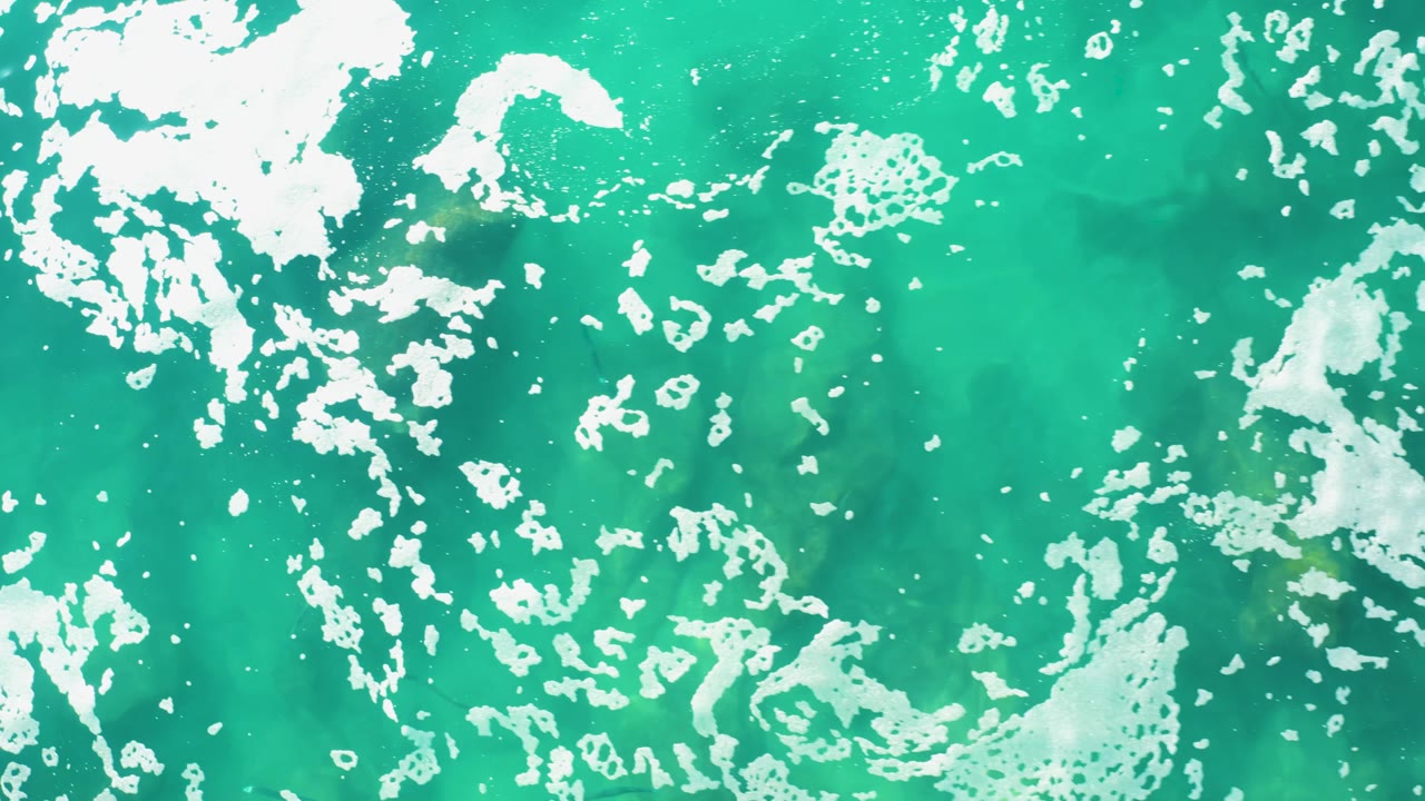 Laguna Beach Los Cabos ocean water viewed from above, with denser white foam crossing the upper left and turquoise water visible beneath, no visible people.
