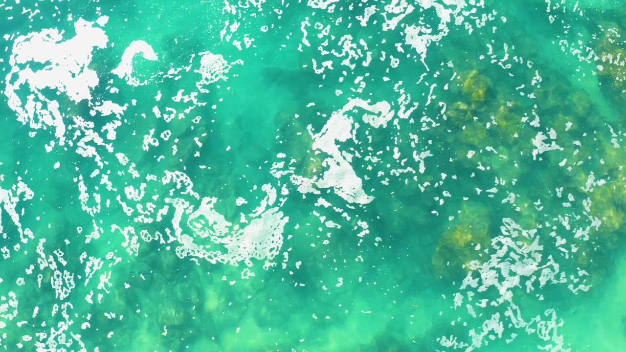 Aerial water detail from Laguna Beach Los Cabos with broken white foam near the upper left and darker submerged areas visible below, with no visible people.