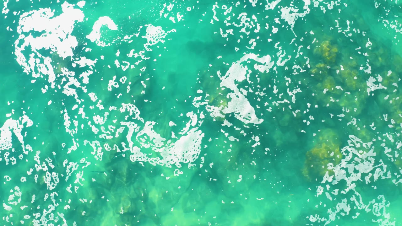 Top-down Laguna Beach Los Cabos water surface with bright white foam streaks crossing the upper and lower frame and no visible people.