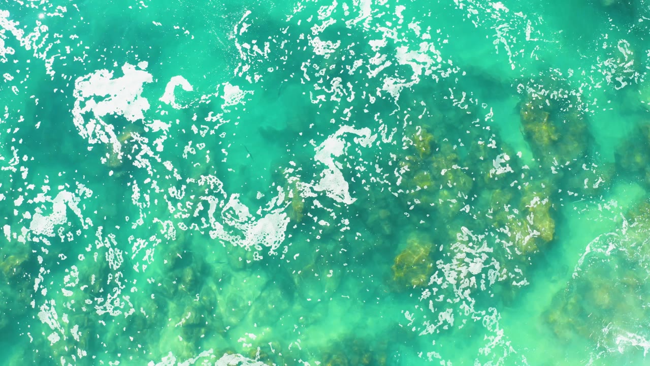 At 0:01, white foam patches sit farther across the turquoise water while darker submerged features remain visible below, with no visible people.