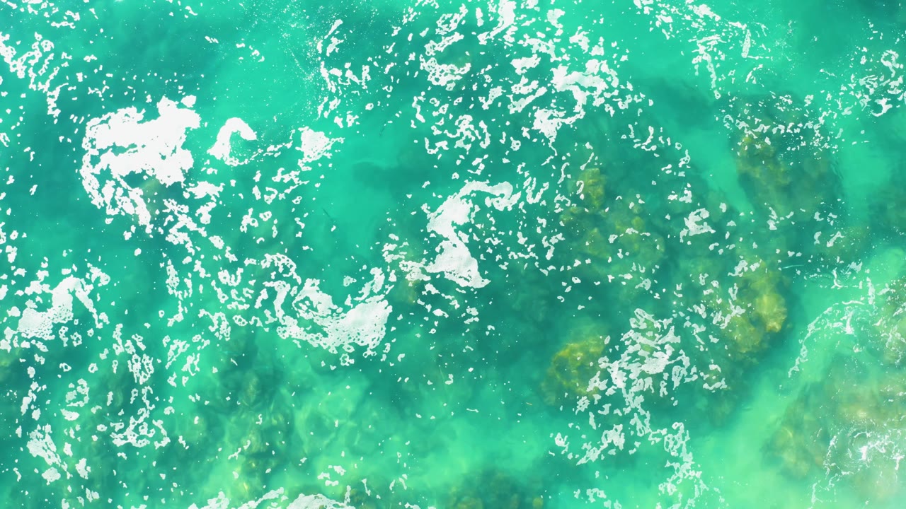 At 0:03, a denser curl of white seafoam crosses the left side of the turquoise ocean surface while submerged rocks remain visible below, with no visible people.