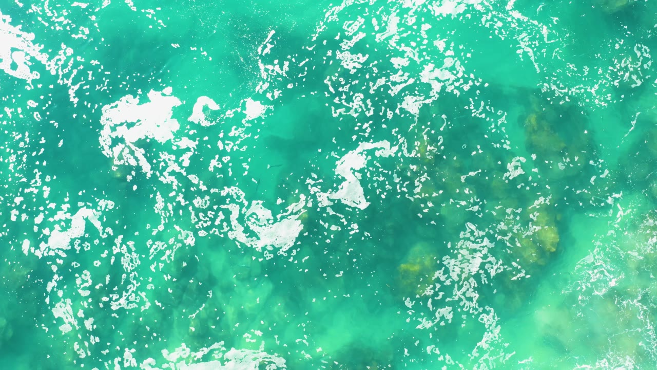 Clear turquoise water fills the frame at Laguna Beach Los Cabos, with white foam streaks crossing the upper half, pale green seabed patches below, and no visible people.
