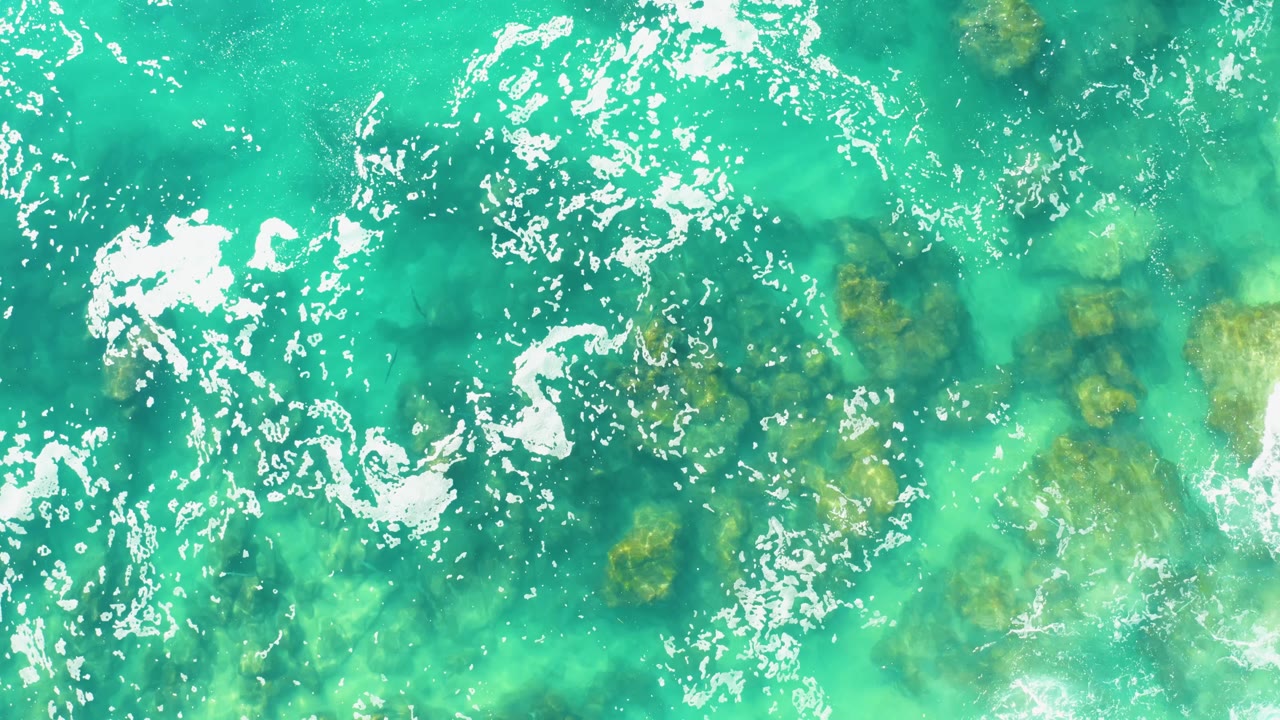 Clear turquoise ocean at Laguna Beach Los Cabos fills the frame, with bright foam patches near the top and darker seabed rocks showing through, no visible people.