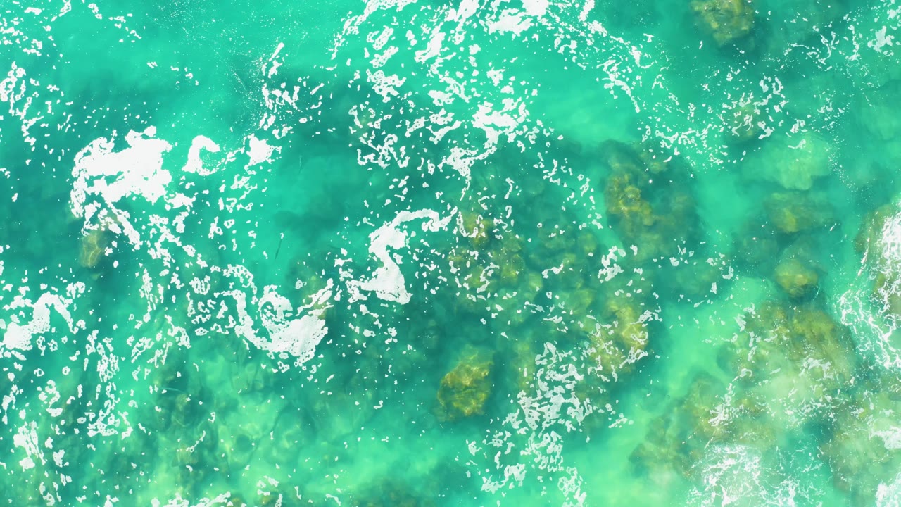 Turquoise water at Laguna Beach Los Cabos shows clustered white foam at left and center, with green-brown underwater rocks visible through the water, no visible people.