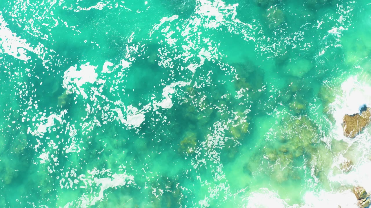 Overhead frame from Laguna Beach Los Cabos showing turquoise water, scattered white foam, a pale foamy area at lower right, and no visible people.