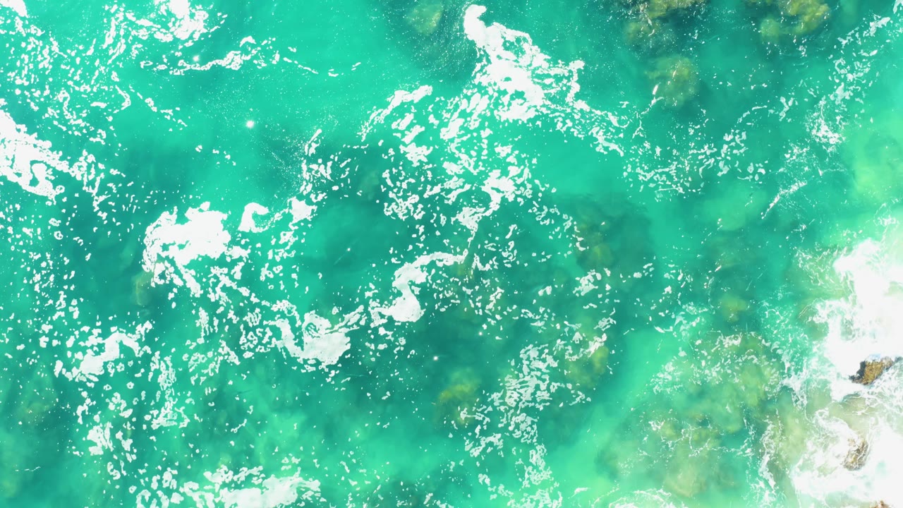 Laguna Beach Los Cabos water seen from overhead with green-blue shallows, white foam sweeping across the top, small rocks on the right side, and no visible people.