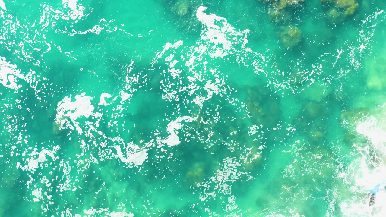Overhead Laguna Beach Los Cabos frame with bright turquoise water, scattered foam clusters, darker submerged rocks, and no visible people.