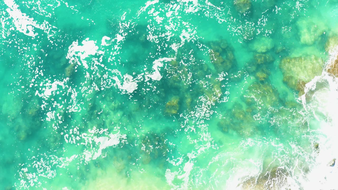 Turquoise and green-blue water at Laguna Beach Los Cabos shows shifting white foam trails above darker submerged rocks with no visible people.