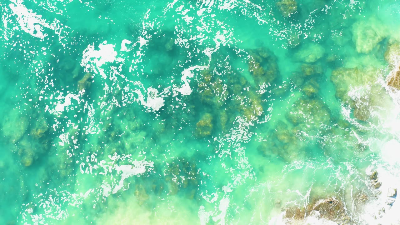 Overhead Laguna Beach Los Cabos water view with white foam bands, darker underwater rocks, and brighter wash beginning along the right edge, with no visible people.