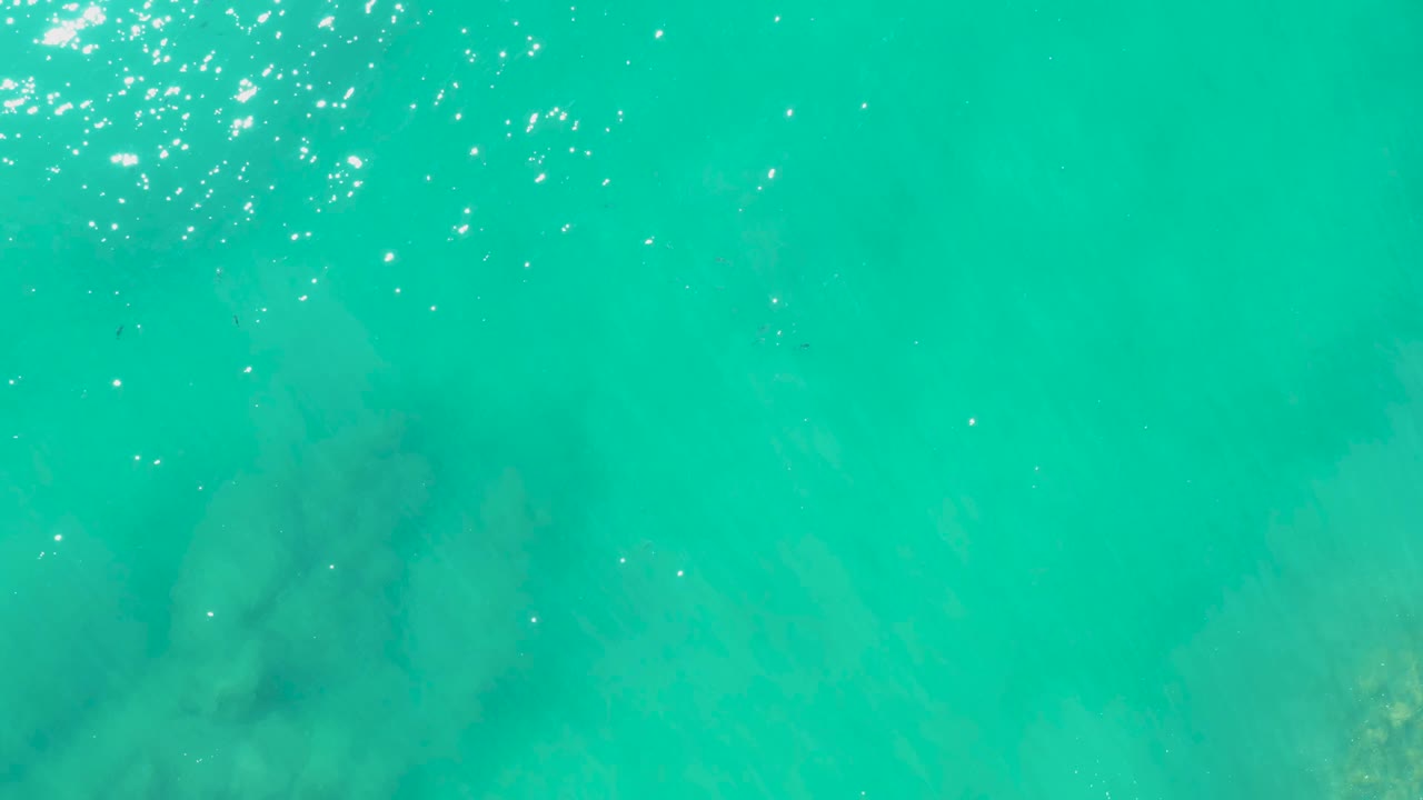 At 0:01, the Laguna Beach Los Cabos water remains a clear teal field with clustered sun glints across the upper edge and soft rock shadows under the surface, with no visible people.