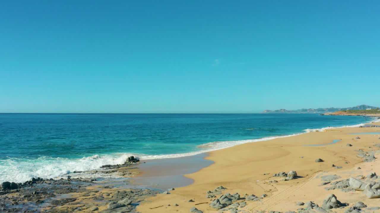 Wide sandy beach with turquoise surf and rocks under clear sky